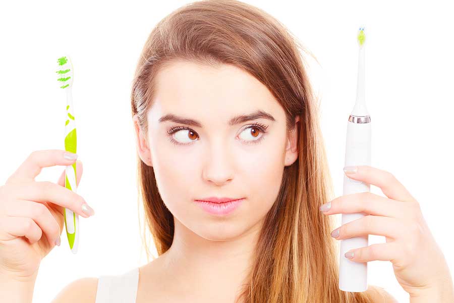 A woman holds a manual toothbrush in one hand and an electric toothbrush in the other hand. She is looking and considering which brush to use.